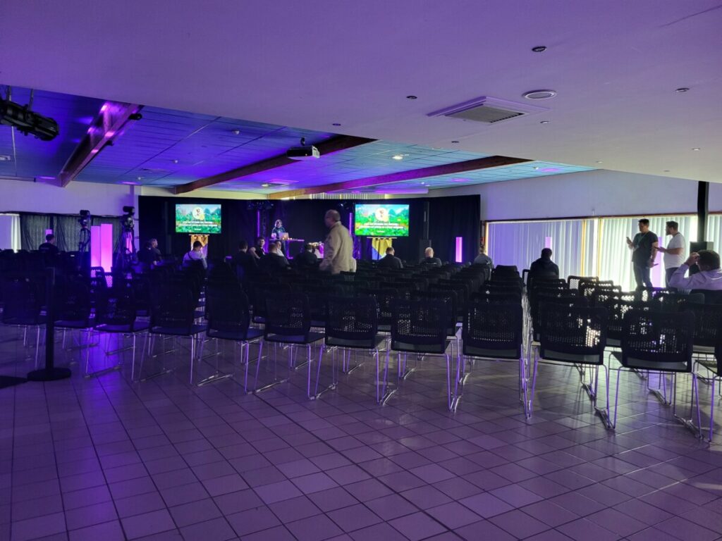Salle de conférence avec des participants et des présentations sur écran. Ambiance sombre et sièges vides.