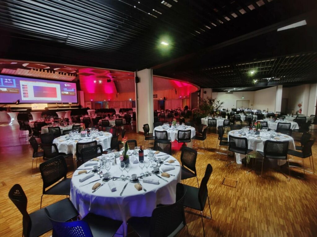 Salle de réception avec tables dressées pour un événement, scène en arrière-plan, lumière ambiante.