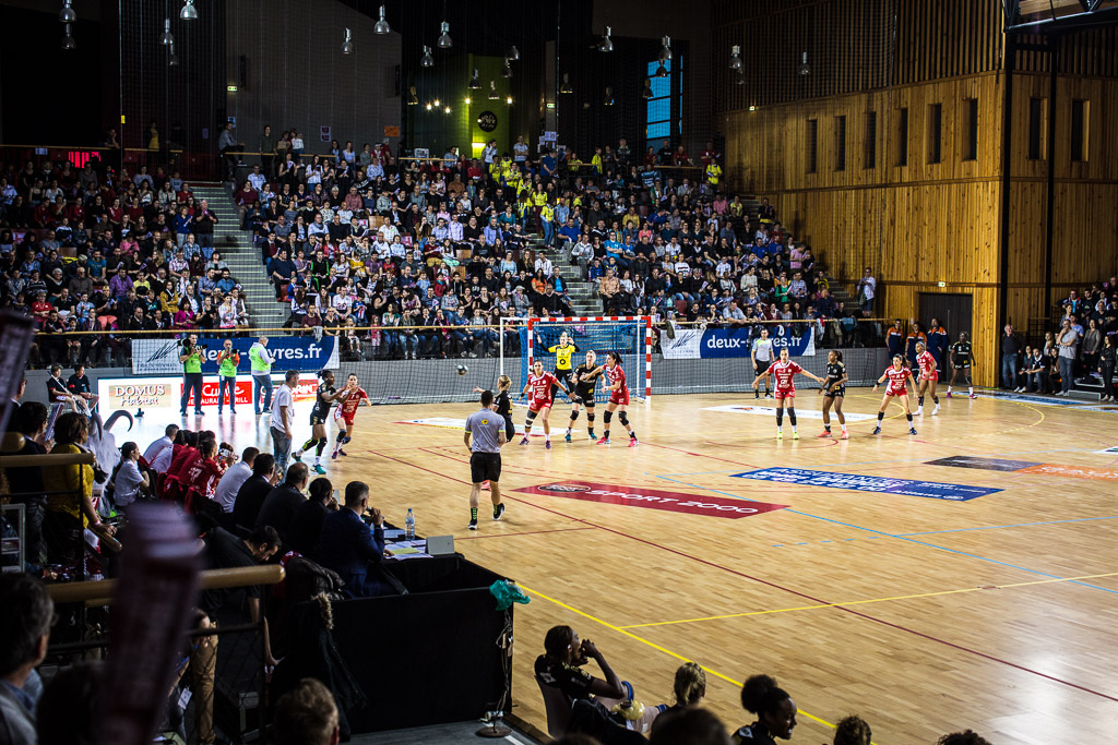 Match de handball en salle avec des joueurs en action et un public nombreux. Atmosphère sportive énergique.