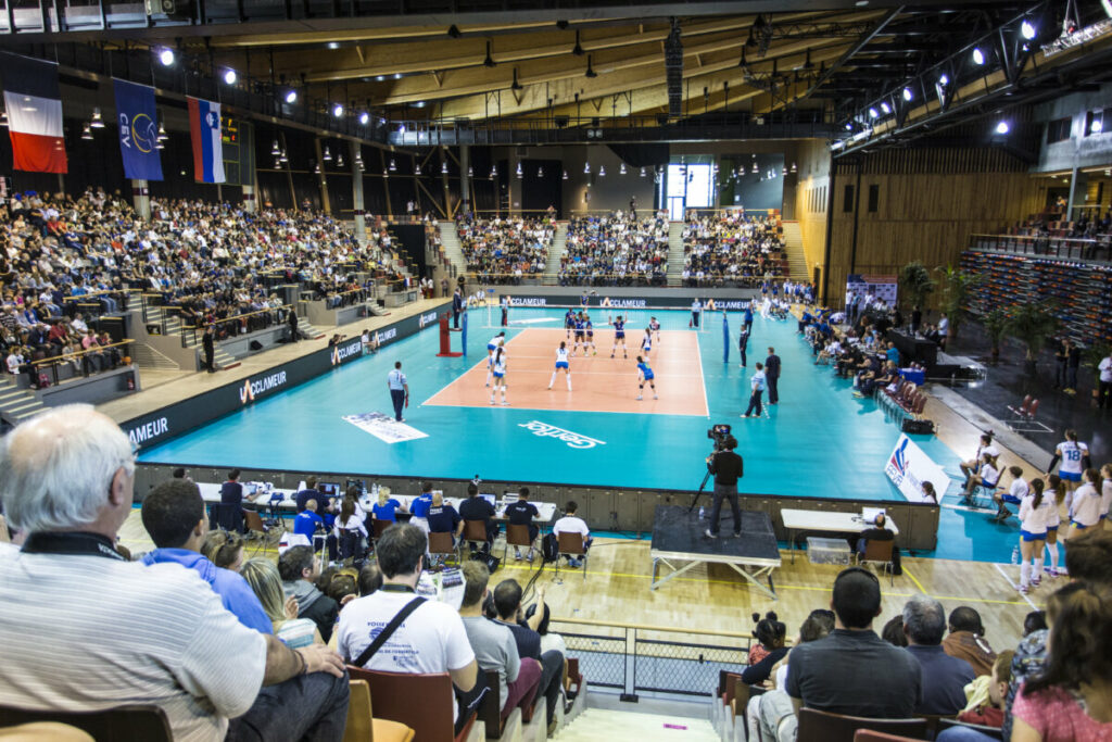 Match de volley-ball dans un gymnase bondé, avec des spectateurs enthousiastes et des équipes en action.