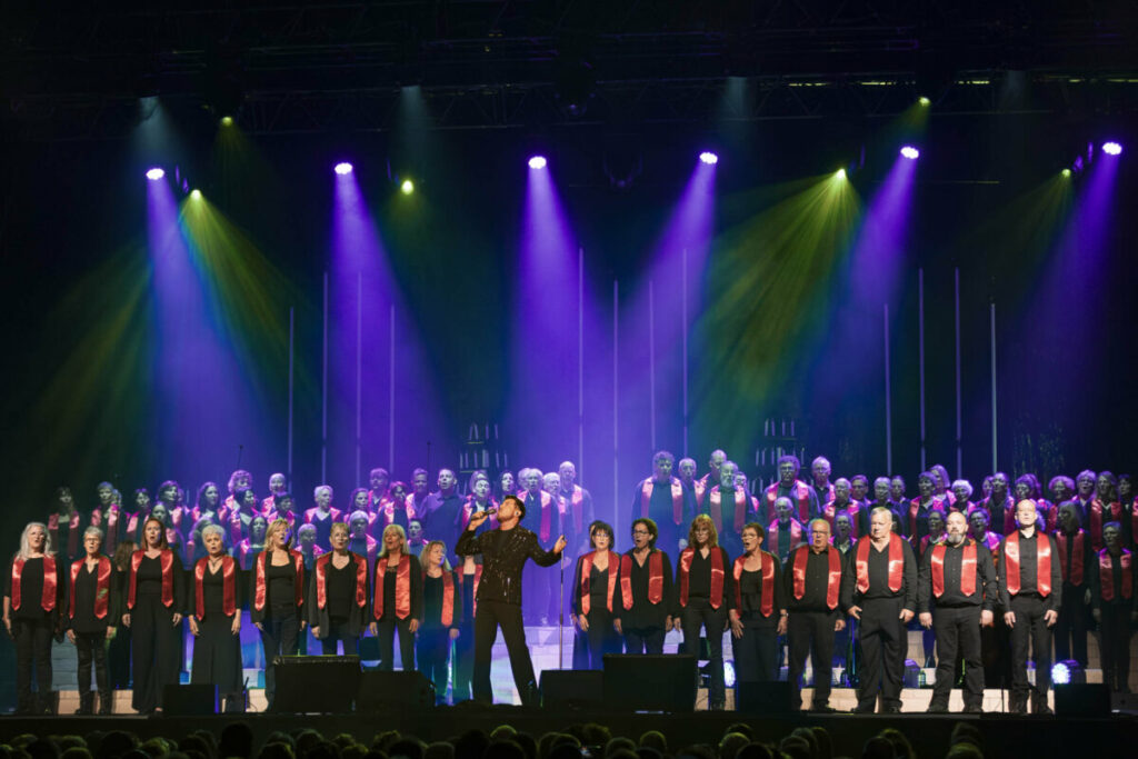 Concert choral avec un soliste en avant et des choristes en arrière-plan, sous des lumières colorées.