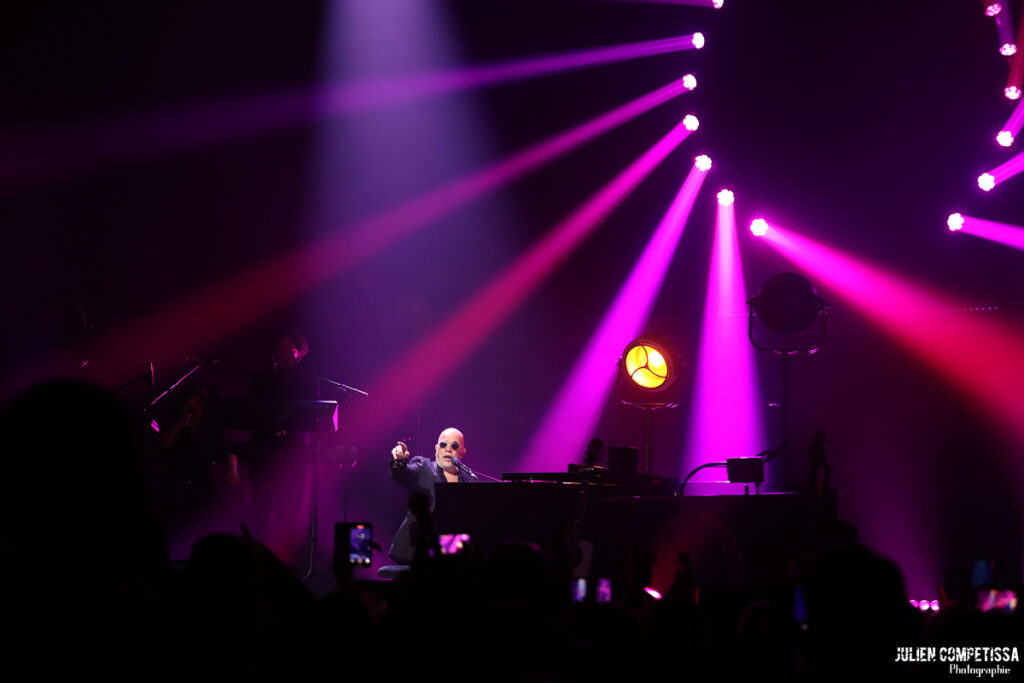 Un artiste sur scène joue du piano pendant un concert, avec des lumières colorées en arrière-plan.