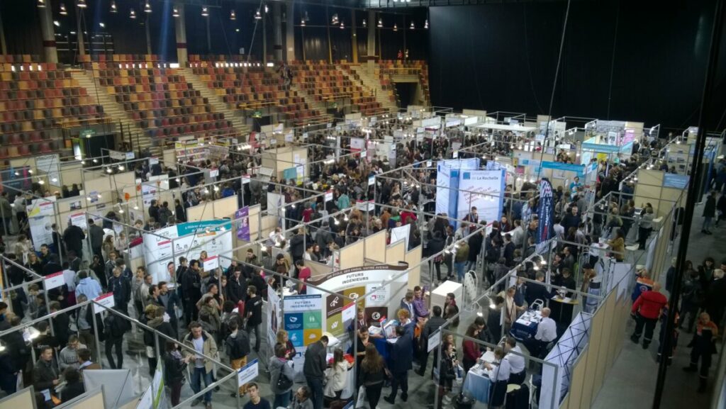 Foule réunie lors d'une foire, interagissant avec divers stands d'exposition.