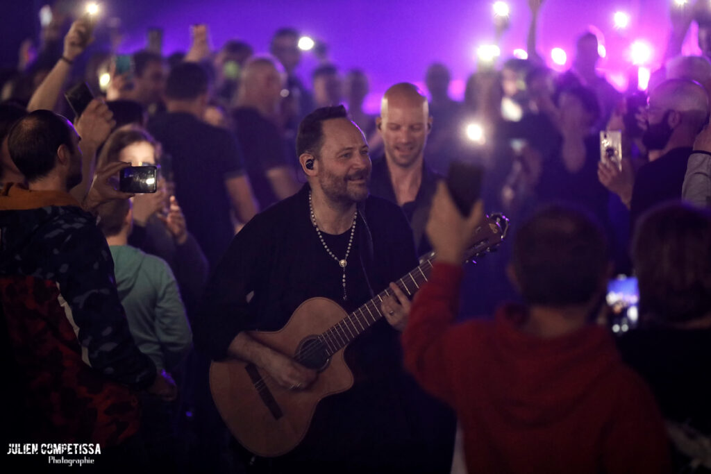Homme jouant de la guitare au milieu d'une foule enthousiaste, éclairée par des lumières de téléphones.