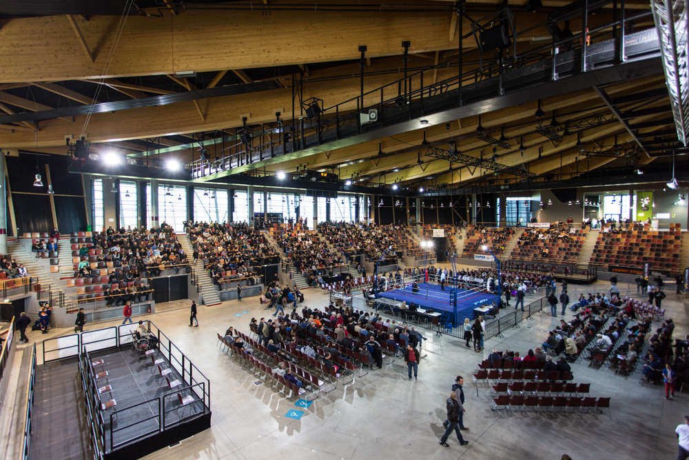 Salle de spectacle remplie de spectateurs, avec un ring de boxe au centre.