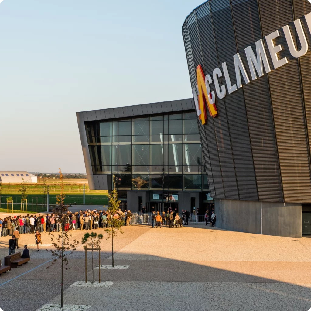 Une foule fait la queue devant un bâtiment moderne, prêt à entrer à l'intérieur lors d'un événement.