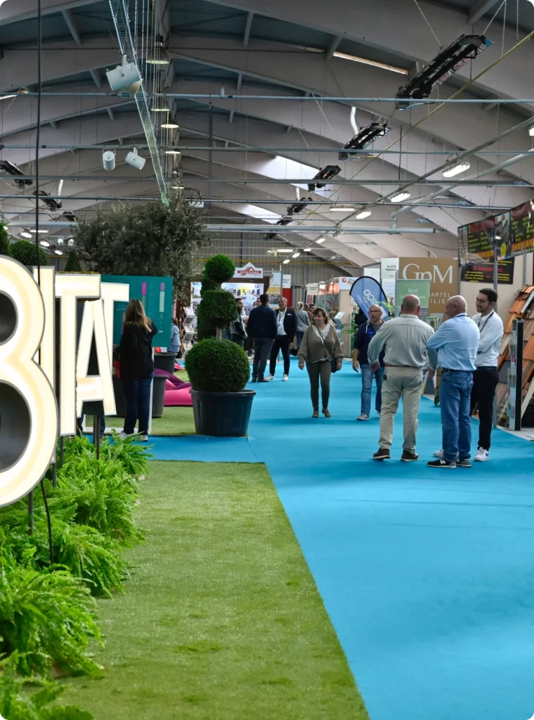 Salon professionnel avec visiteurs interagissant et des stands exposants, allée centrale décorée de verdure.