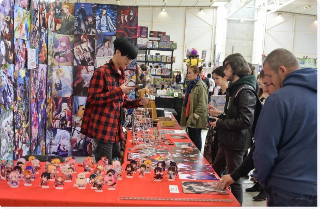 Stand de vente animé avec des personnes examinant des figurines et des affiches, discutant et découvrant des produits.
