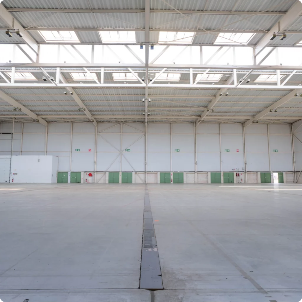 Un vaste espace intérieur avec de grandes fenêtres et plusieurs portes vertes. L'endroit est bien éclairé et vide.