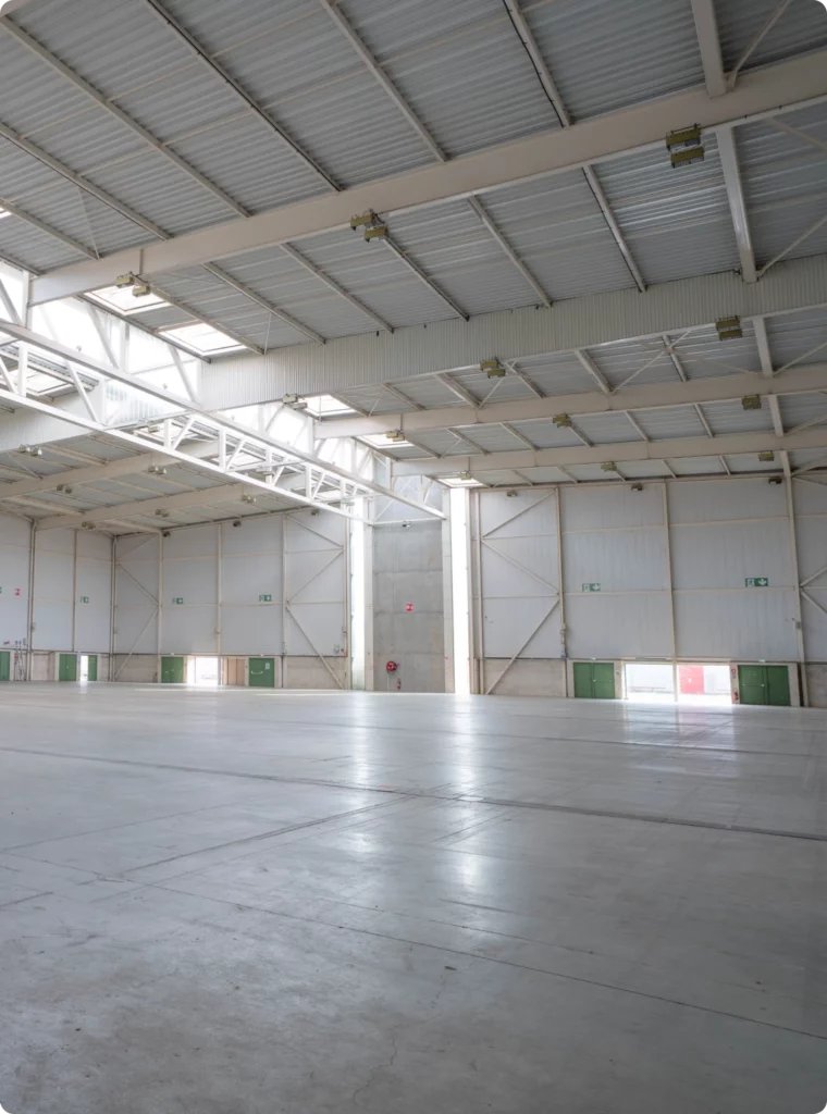 Grande salle vide avec murs en métal, fenêtres sur le plafond et portes d'accès. Espace pour événements ou entreposage.