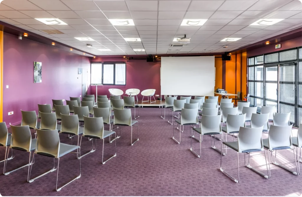 Salle de réunion avec des chaises disposées pour un événement, écran de projection à l'avant.