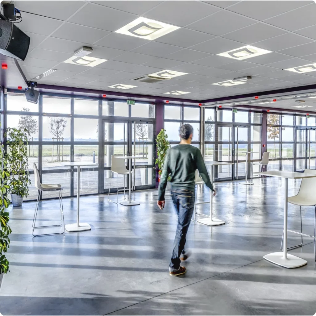 Un homme marche dans un espace lumineux avec des tables et des plantes, près de grandes fenêtres.