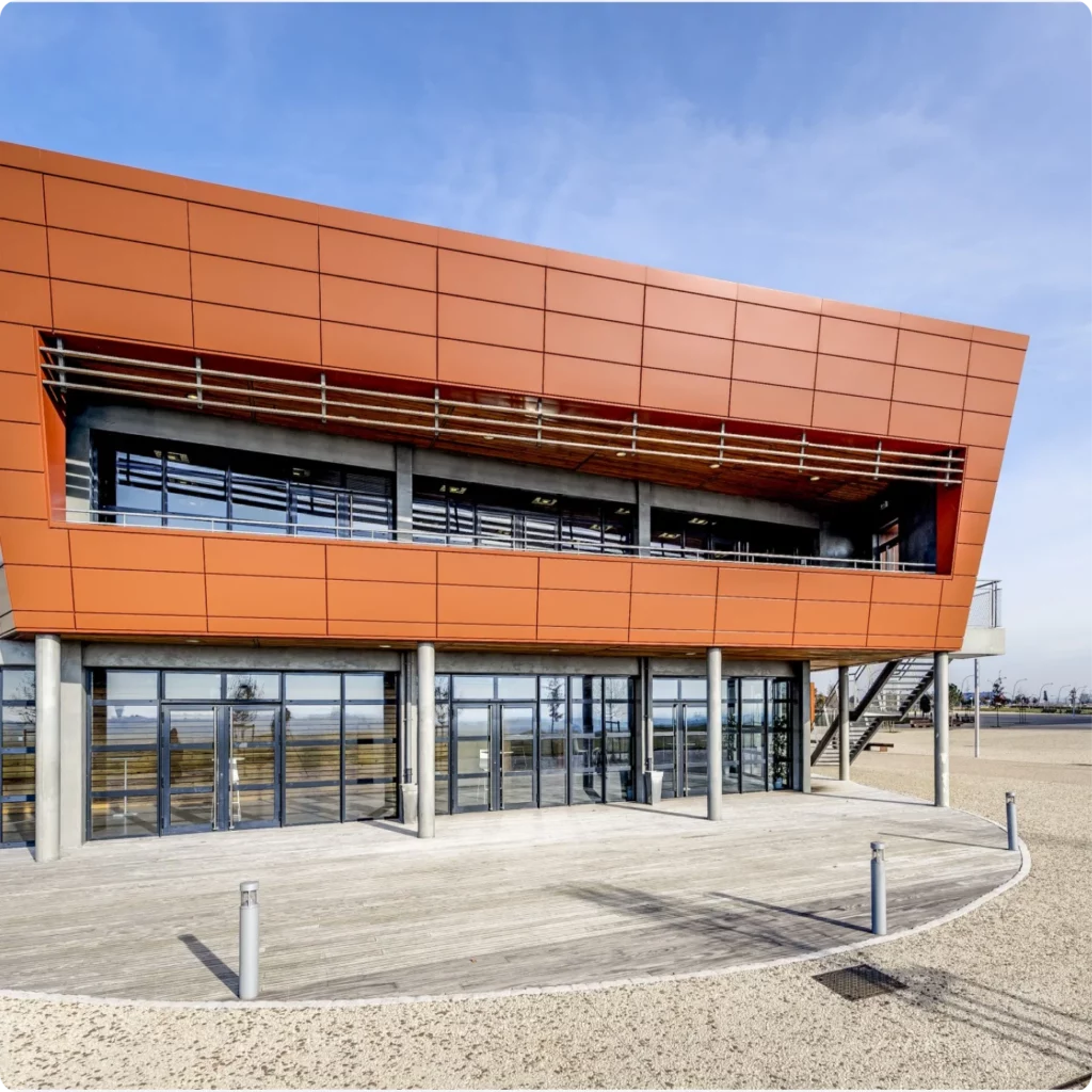 Bâtiment moderne avec façade en panneaux orange et large baie vitrée, situé près d'une zone extérieure aménagée.