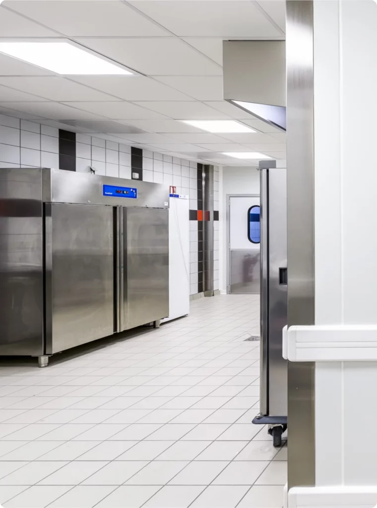 Cuisine moderne avec réfrigérateurs en inox, murs carrelés et porte menant à un espace de travail.