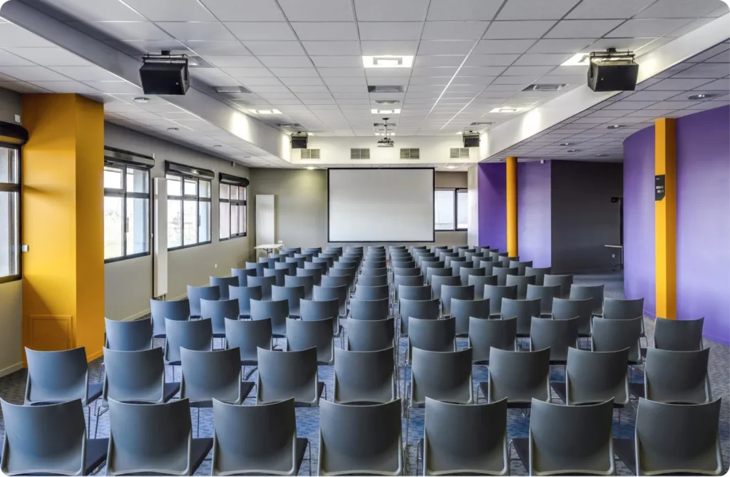 Salle de conférence avec rangées de chaises et un écran de projection au fond.