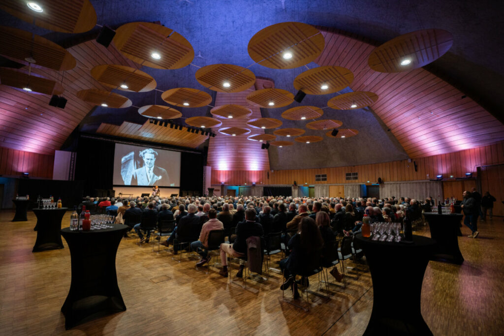 Une foule assiste à une conférence dans une salle moderne avec une grande scène et un écran projetant des images.